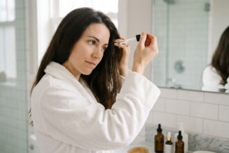 Femme appliquant un sérum anti-chute sur le cuir chevelu dans une salle de bain moderne avec des flacons de soins capillaires sur le comptoir